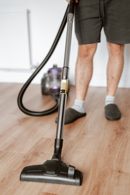 Close-up of a person using a handheld vacuum cleaner on a light wooden floor in a modern living room, with the person's legs visible wearing dark shorts and socks. The vacuum has a black and grey body