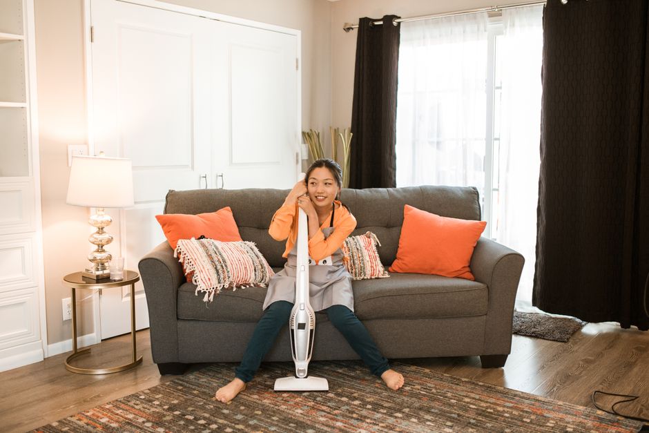 Close-up of a person using a handheld vacuum cleaner on a light wooden floor in a modern living room, with the person's legs visible wearing dark shorts and socks. The vacuum has a black and grey body