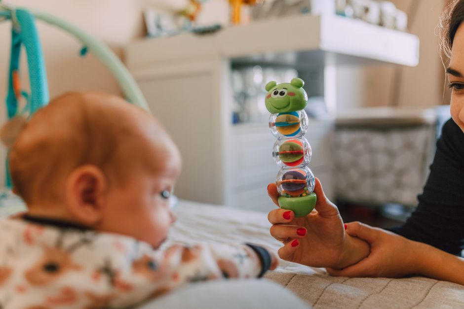 A caregiver is sitting on a bed in a warmly lit bedroom, holding a stack of colorful plastic stacking toys, including a green frog at the top, in front of a young child with light red hair wearing a p