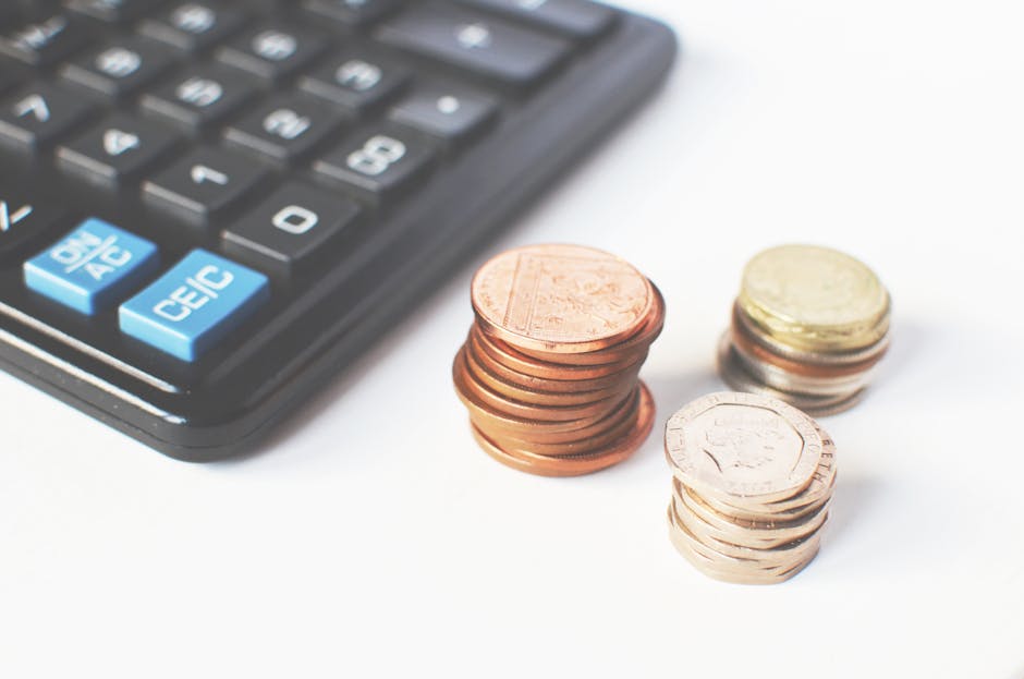 Close-up image of a black calculator with blue and white buttons, positioned on the left side of the frame, alongside several neatly stacked coins of various denominations and metallic colors on the r