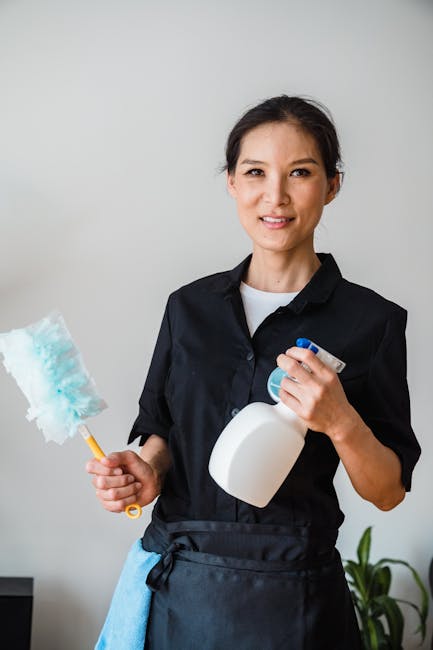 A professional cleaner from cleaninghousecleaners.com stands indoors holding a spray bottle of cleaning solution in one hand and a colorful duster in the other. She is dressed in a black uniform with 